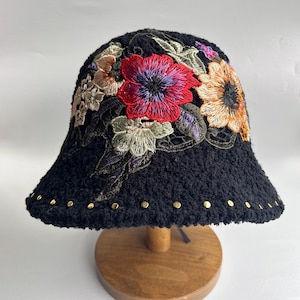 Puede incluir: Sombrero cubo negro con flores bordadas en tonos rojos, naranjas, morados y verdes. El sombrero tiene una textura peluda y detalles dorados alrededor del borde.