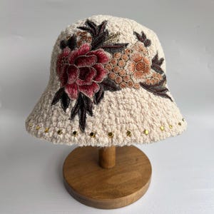 May include: A white crocheted bucket hat with a floral embroidery design. The embroidery features a large pink flower with smaller yellow and orange flowers and green leaves. The hat has gold studs around the brim.