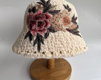 Puede incluir: Un sombrero de cubo de crochet blanco con un diseño de bordado floral. El bordado presenta una gran flor rosa con flores más pequeñas de color amarillo y naranja y hojas verdes. El sombrero tiene tachuelas doradas alrededor del borde.