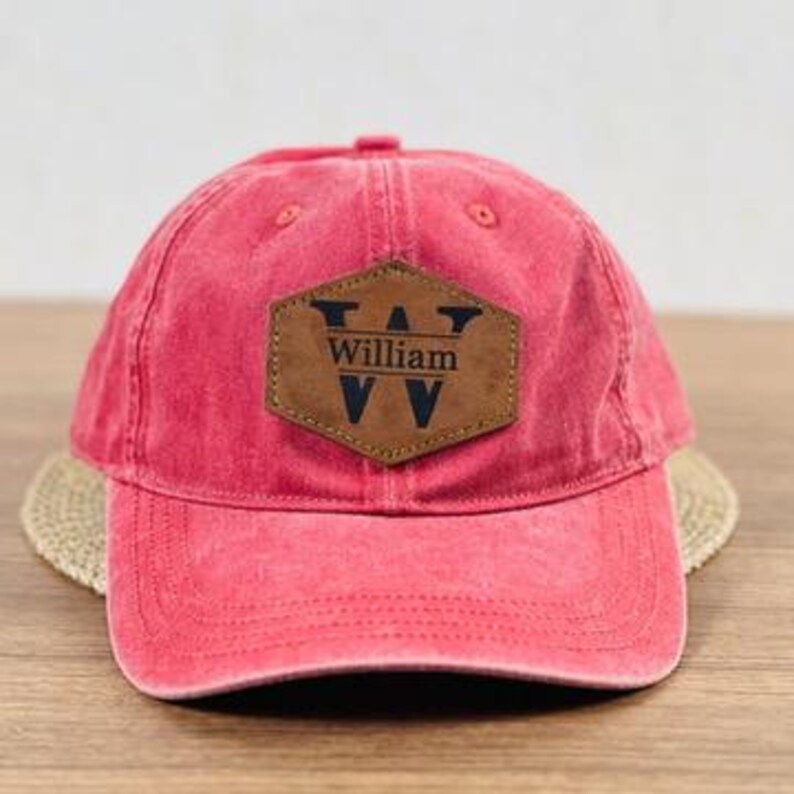 May include: A red baseball cap with a brown leather patch. The patch is in the shape of a hexagon and has the name "William" and the letter "W" printed on it. The cap has a curved brim and an adjustable strap.
