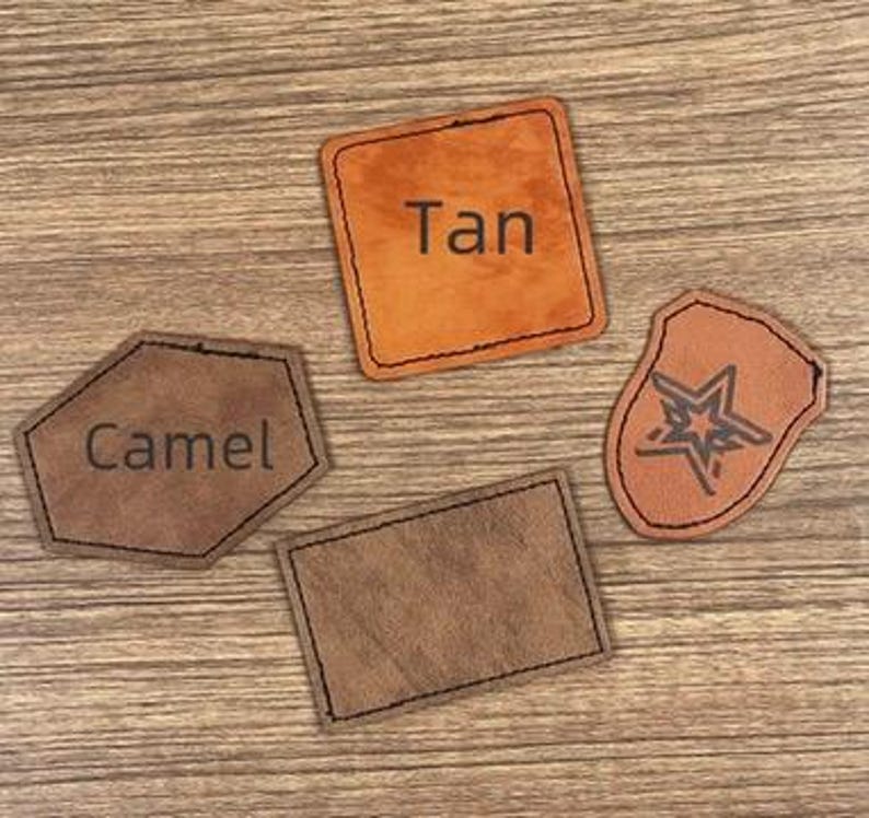 May include: Four leather patches in shades of brown and tan are arranged on a wooden surface. One patch is a rectangle, another is a square with "Tan" printed on it, a third is a shield shape with a star design, and the last is a hexagon with "Camel" printed on it.