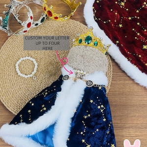 May include: A royal pet costume set featuring a navy blue cape with gold stars and white faux fur trim, a gold crown with green jewels, and a matching pearl bracelet. The text reads "CUSTOM YOUR LETTER UP TO FOUR HERE".