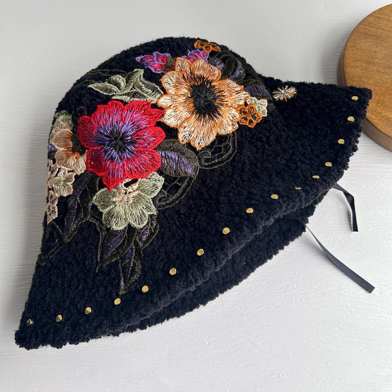 Puede incluir: Sombrero de cubo negro y peludo con flores bordadas en tonos rojos, naranjas, amarillos y verdes. El sombrero tiene tachuelas doradas alrededor del borde.