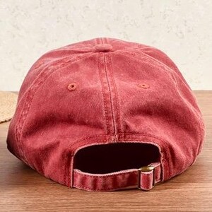 May include: A faded red baseball cap with a curved brim and adjustable metal buckle closure. The cap is made of a soft, worn material, and the back features an opening for a ponytail. The hat is sitting on a wooden surface.