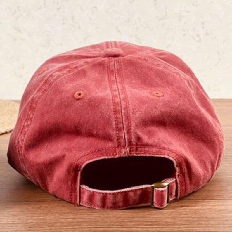 May include: A faded red baseball cap with a curved brim and adjustable metal buckle closure. The cap is made of a soft, worn material, and the back features an opening for a ponytail. The hat is sitting on a wooden surface.