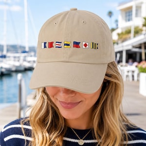 May include: A beige baseball cap featuring a horizontal row of seven colourful nautical signal flags embroidered across the front. The cap has a structured crown, a curved brim, and stitched eyelets for ventilation, providing a classic look.