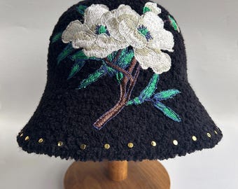 Puede incluir: Sombrero negro de cubo con un diseño de flores blancas bordadas. El sombrero tiene tachuelas doradas alrededor del borde.