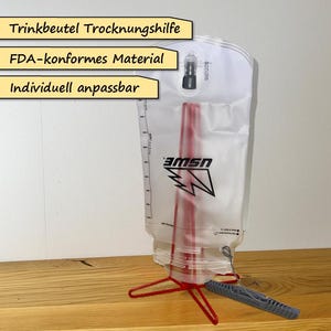 Op de afbeelding: Een doorzichtige drinkreservoir met een rode rietje en een zwart logo. Het reservoir wordt ondersteund door een rode standaard en heeft een grijze bijtklep. Tekst op de afbeelding is "Trinkbeutel Trocknungshilfe", "FDA-konformes Material" en "Individuell anpassbar".
