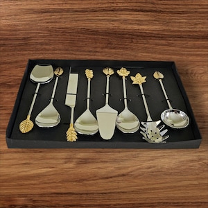 May include: A set of eight silver serving utensils with gold leaf accents. The utensils include a cake server, a spoon, a ladle, a fork, and a slotted spoon. The utensils are arranged in a black box.