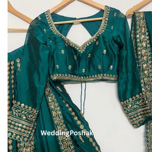 K&ouml;nnte beinhalten: Smaragdgr&uuml;nes formelles Outfit mit goldener Stickerei. Das Set beinhaltet ein lang&auml;rmliges Oberteil mit V-Ausschnitt und einen passenden Rock. Der Text "Wedding Poshak" ist auf dem Rock zu sehen.