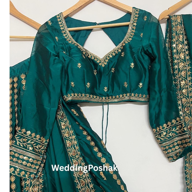 K&ouml;nnte beinhalten: Smaragdgr&uuml;nes formelles Outfit mit goldener Stickerei. Das Set beinhaltet ein lang&auml;rmliges Oberteil mit V-Ausschnitt und einen passenden Rock. Der Text "Wedding Poshak" ist auf dem Rock zu sehen.
