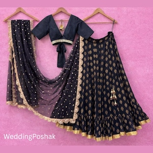 以下が含まれることがあります： ピンク色の背景に、黒と金のインドのウェディング衣装が吊り下げられています。トップス、スカート、スカーフが含まれています。スカートとスカーフには金のトリムが付いています。トップスは黒で、深いVネックです。WeddingPoshakから。