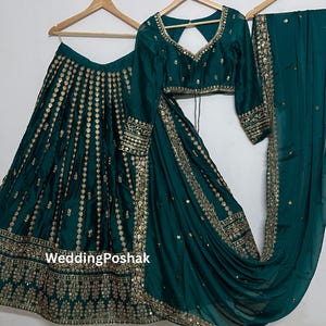 K&ouml;nnte beinhalten: Ein tealgr&uuml;nes indisches Hochzeitsoutfit, bestehend aus Rock, Bluse und Schal, allesamt mit goldener Stickerei und Verzierungen verziert. Das Outfit wird auf Holzb&uuml;geln pr&auml;sentiert.