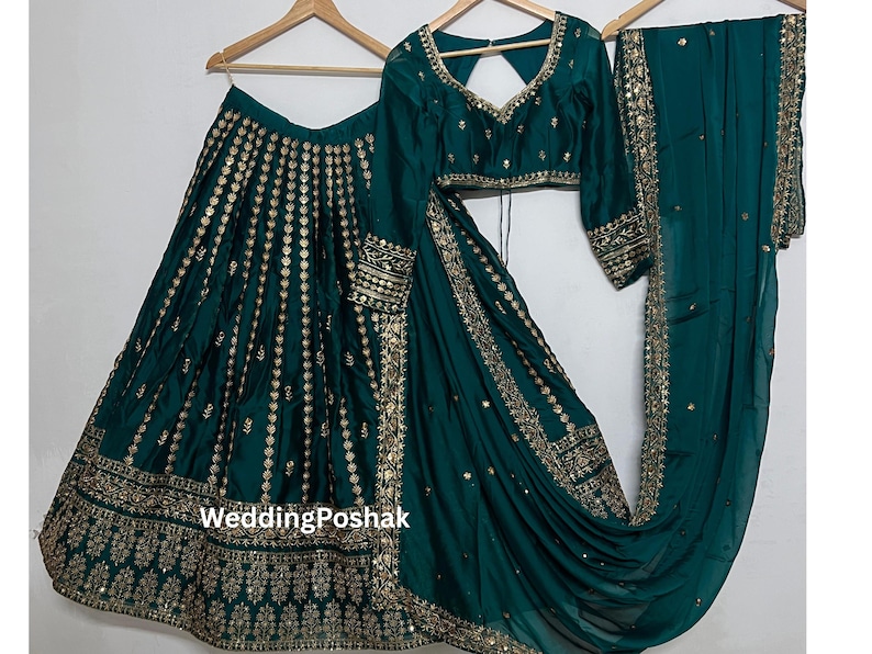 K&ouml;nnte beinhalten: Ein tealgr&uuml;nes indisches Hochzeitsoutfit, bestehend aus Rock, Bluse und Schal, allesamt mit goldener Stickerei und Verzierungen verziert. Das Outfit wird auf Holzb&uuml;geln pr&auml;sentiert.