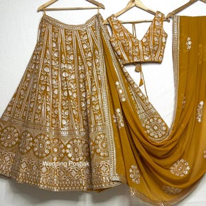 Puede incluir: Conjunto lehenga bordado de color mostaza con blusa y dupatta a juego. El lehenga presenta intrincados patrones florales y geométricos en hilo dorado. La blusa tiene cuello en V y espalda anudada. El dupatta está drapeado sobre el conjunto.