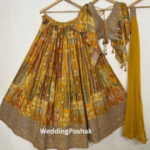 Puede incluir: Un conjunto de tres piezas amarillo mostaza con una falda estampada, un top corto y una bufanda a juego. La falda y el top presentan un diseño detallado con dorado y otros colores. El top tiene cuello en V y mangas cortas. El texto "WeddingPoshak" es visible.