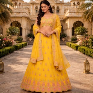 May include: A woman in a bright yellow lehenga choli, a traditional Indian outfit. The set includes a crop top, a long skirt, and a matching scarf. The outfit is decorated with detailed embroidery in pink and gold. The background shows a palace and manicured gardens.