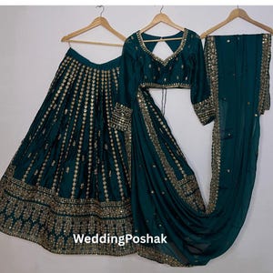 K&ouml;nnte beinhalten: Ein dreiteiliges formelles Outfit in Teal, bestehend aus einem Rock, einem Oberteil mit langen &Auml;rmeln und einem drapierten Schal. Rock und Oberteil sind mit goldenen Verzierungen versehen. Das Outfit wird auf Holzb&uuml;geln vor wei&szlig;em Hintergrund pr&auml;sentiert. Der Text "WeddingPoshak" ist sichtbar.