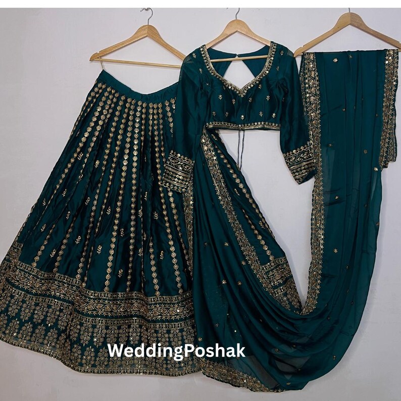 K&ouml;nnte beinhalten: Ein dreiteiliges formelles Outfit in Teal, bestehend aus einem Rock, einem Oberteil mit langen &Auml;rmeln und einem drapierten Schal. Rock und Oberteil sind mit goldenen Verzierungen versehen. Das Outfit wird auf Holzb&uuml;geln vor wei&szlig;em Hintergrund pr&auml;sentiert. Der Text "WeddingPoshak" ist sichtbar.