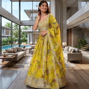 Op de afbeelding: Een vrouw draagt een gele lehenga choli met bloemenprint. De outfit bestaat uit een crop top, een lange rok en een bijpassende dupatta. De lehenga heeft een gouden rand en een bloemenpatroon. De achtergrond toont een modern huis met grote ramen.