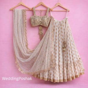May include: A cream-coloured bridal outfit with a gold-embellished bodice, a matching skirt with gold detailing, and a sheer dupatta. The outfit is displayed on wooden hangers against a pink backdrop. The text "Wedding Poshak" is visible.