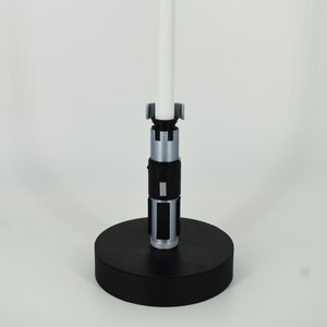 K&ouml;nnte beinhalten: Ein Lichtschwert-Replikat mit wei&szlig;er Klinge und silbernem und schwarzem Griff. Das Lichtschwert ist auf einem schwarzen, kreisf&ouml;rmigen Sockel montiert und steht aufrecht vor einem wei&szlig;en Hintergrund. Das Gesamtdesign ist elegant und detailliert.