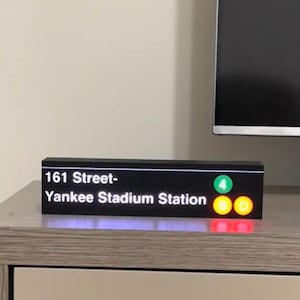 May include: A black rectangular sign with the text "161 Street-Yankee Stadium Station" in white. The sign has a green circle with the number 4, and two orange lights. The sign is on a wooden surface.