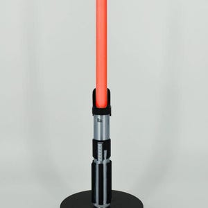Op de afbeelding: Een zwart lightsaber met een rode blade op een zwarte basis. De lightsaber heeft een zilveren handvat met zwarte accenten.