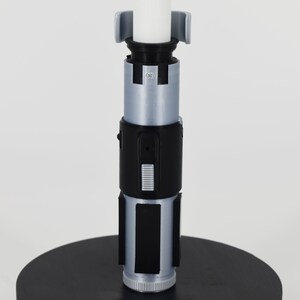 K&ouml;nnte beinhalten: Ein detailgetreues Lichtschwert mit wei&szlig;er Klinge und einem schwarzen und silbernen Griff. Der Griff hat rechteckige und zylindrische Komponenten und ist auf einem schwarzen kreisf&ouml;rmigen Sockel montiert. Das Lichtschwert ist ein Sammlerst&uuml;ck.