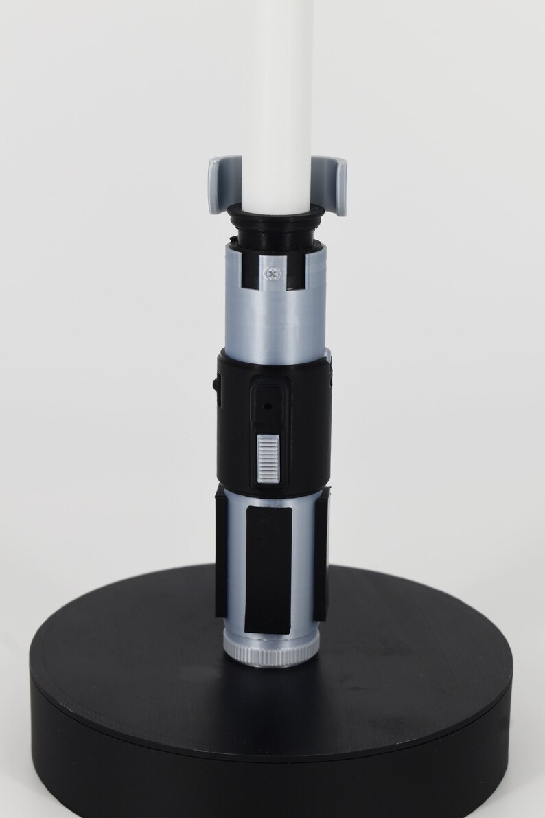 K&ouml;nnte beinhalten: Ein detailgetreues Lichtschwert mit wei&szlig;er Klinge und einem schwarzen und silbernen Griff. Der Griff hat rechteckige und zylindrische Komponenten und ist auf einem schwarzen kreisf&ouml;rmigen Sockel montiert. Das Lichtschwert ist ein Sammlerst&uuml;ck.