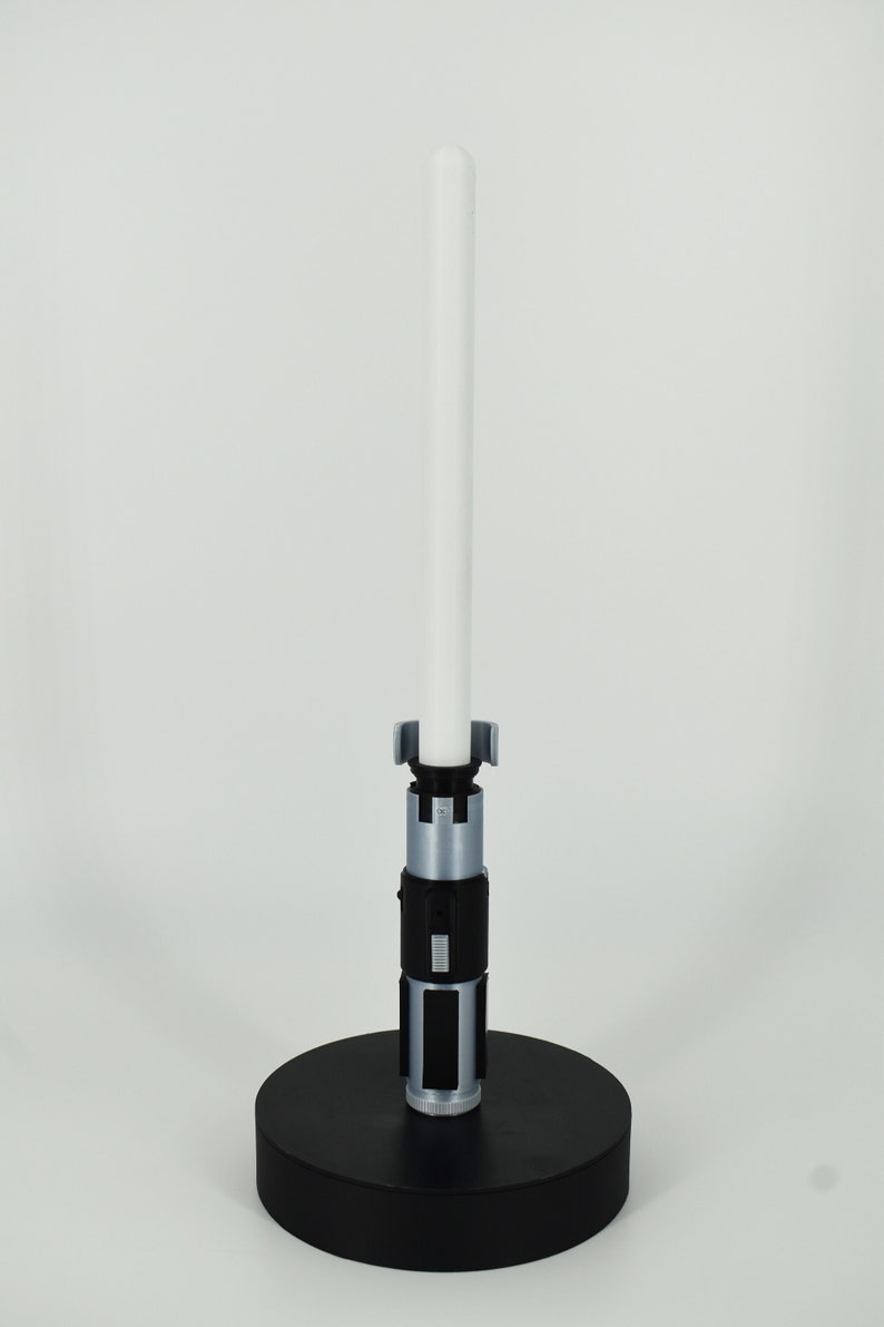 K&ouml;nnte beinhalten: Ein Lichtschwert-Replikat mit wei&szlig;er Klinge und silbernem und schwarzem Griff. Das Lichtschwert ist auf einem schwarzen, kreisf&ouml;rmigen Sockel montiert und steht aufrecht vor einem wei&szlig;en Hintergrund. Das Gesamtdesign ist elegant und detailliert.