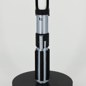 Op de afbeelding: Een zwarte lightsaber-standaard met een zilveren lightsaber erop. De lightsaber heeft een zwarte handgreep met een zilveren blad.