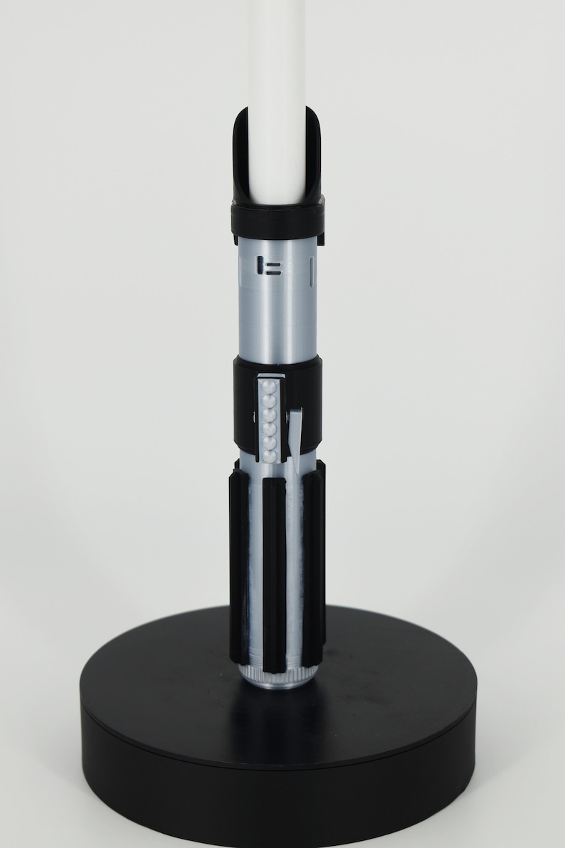 Op de afbeelding: Een zwarte lightsaber-standaard met een zilveren lightsaber erop. De lightsaber heeft een zwarte handgreep met een zilveren blad.