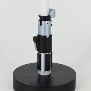 K&ouml;nnte beinhalten: Ein 3D-gedrucktes Lichtschwert-Replikat mit einer wei&szlig;en Klinge und einem detaillierten Griff. Der Griff verf&uuml;gt &uuml;ber silberne und schwarze Komponenten, darunter ein Bedienfeld und rechteckige Akzente. Das Lichtschwert ist auf einer schwarzen kreisf&ouml;rmigen Basis montiert.