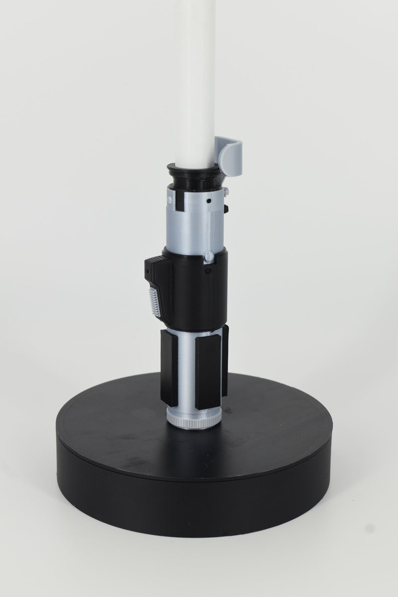 K&ouml;nnte beinhalten: Ein 3D-gedrucktes Lichtschwert-Replikat mit einer wei&szlig;en Klinge und einem detaillierten Griff. Der Griff verf&uuml;gt &uuml;ber silberne und schwarze Komponenten, darunter ein Bedienfeld und rechteckige Akzente. Das Lichtschwert ist auf einer schwarzen kreisf&ouml;rmigen Basis montiert.