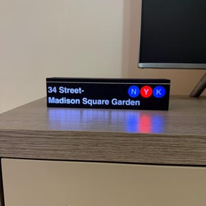 May include: A black rectangular sign with the text "34 Street-Madison Square Garden" in white. The sign also features the letters "NYK" in blue, red, and blue. The sign is sitting on a wooden surface.