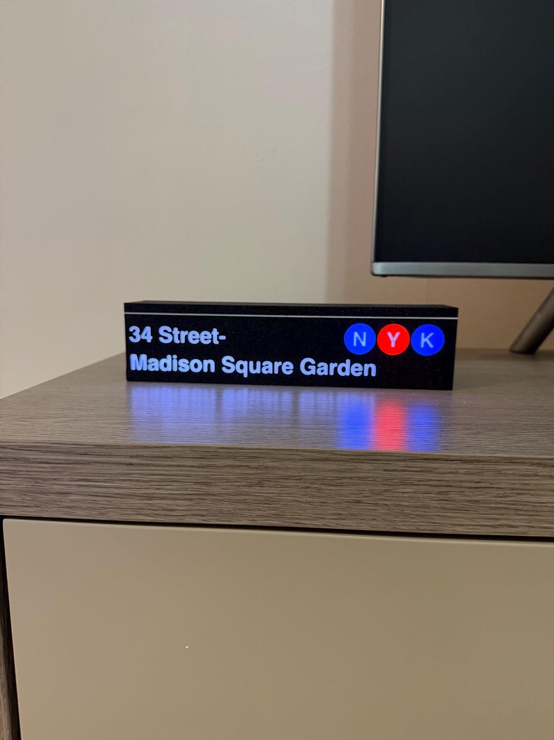 May include: A black rectangular sign with the text "34 Street-Madison Square Garden" in white. The sign also features the letters "NYK" in blue, red, and blue. The sign is sitting on a wooden surface.