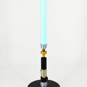 K&ouml;nnte beinhalten: Eine blaue Lichtschwert-Replik mit goldenem Griff und schwarzem Sockel. Das Lichtschwert steht auf einem wei&szlig;en Hintergrund.