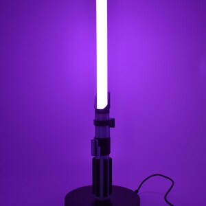 Op de afbeelding: Een paarse lightsaber-lamp met een zwarte voet en een snoer. De lightsaber is aan en de blade is wit.