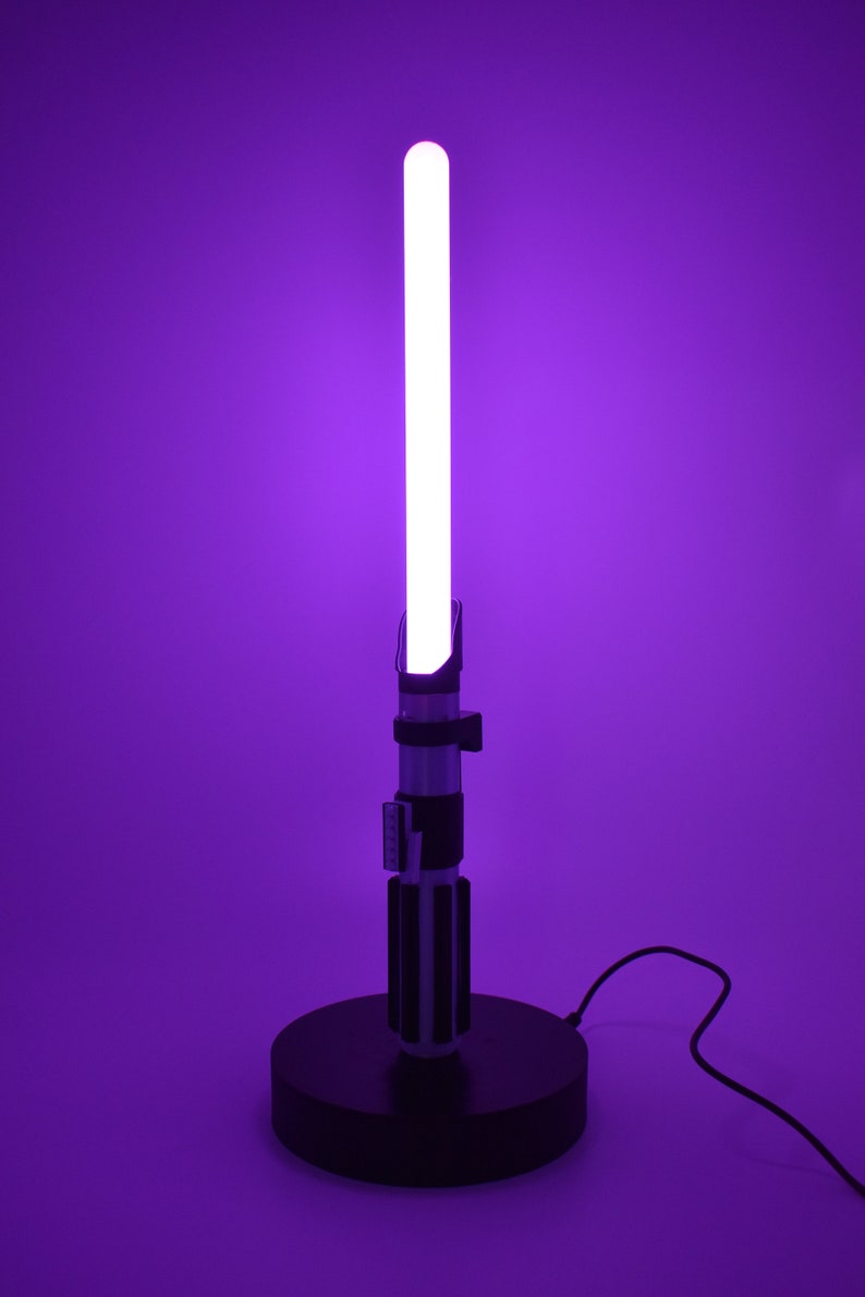 Op de afbeelding: Een paarse lightsaber-lamp met een zwarte voet en een snoer. De lightsaber is aan en de blade is wit.