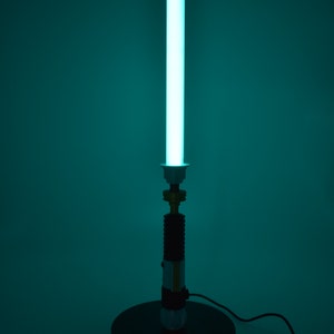 K&ouml;nnte beinhalten: Eine tealblaue Lichtschwertlampe mit schwarzem Sockel und goldener Akzent. Die Lampe ist eingeschaltet und das Licht erhellt den Raum.