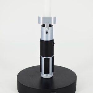 K&ouml;nnte beinhalten: Eine silberne und schwarze Lampe im Lichtschwert-Stil mit wei&szlig;em Licht. Die Lampe hat einen schwarzen, kreisf&ouml;rmigen Sockel mit Ein-/Ausschalter und Stromanschluss. Der Lichtschwertgriff hat schwarze und silberne Details.