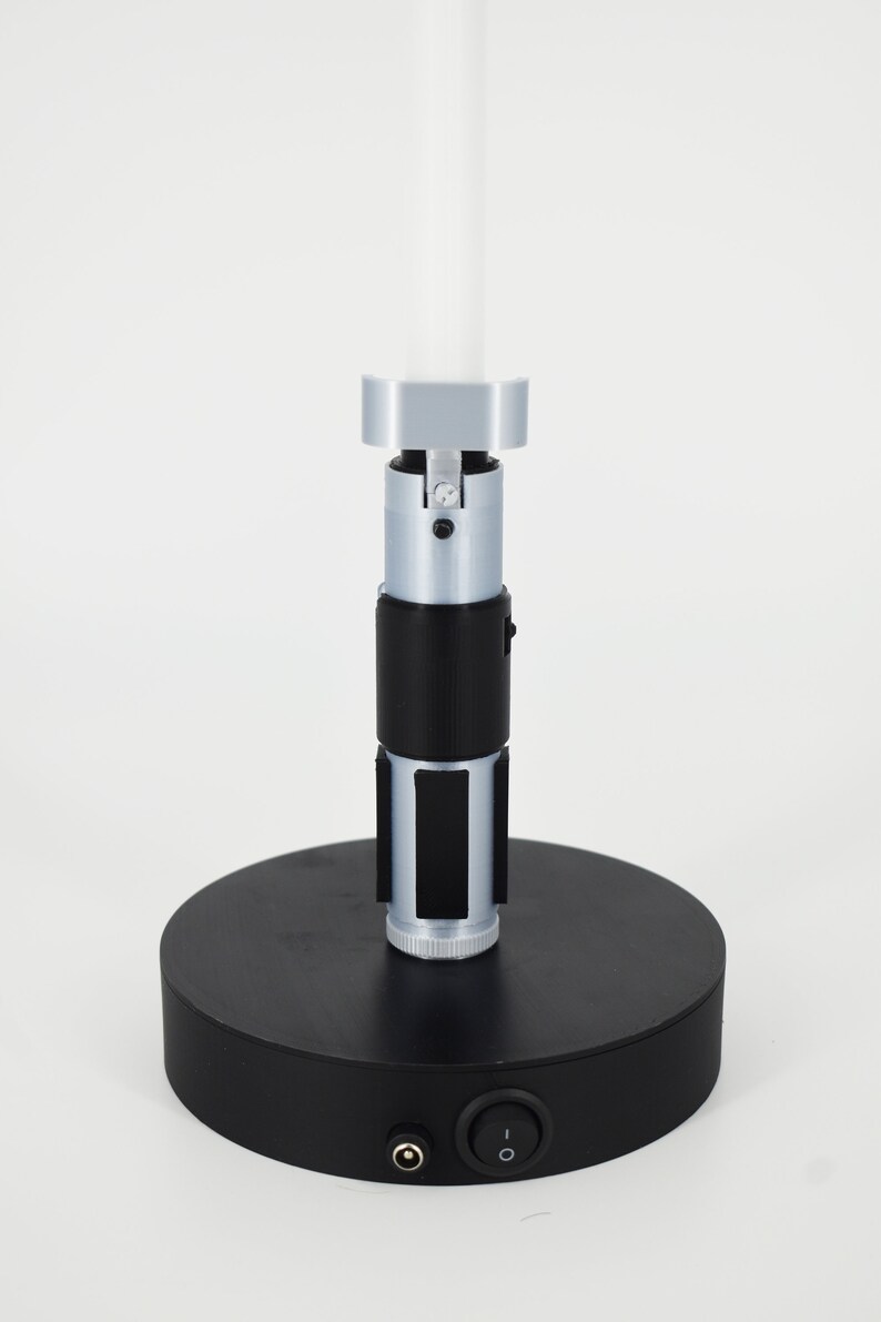 K&ouml;nnte beinhalten: Eine silberne und schwarze Lampe im Lichtschwert-Stil mit wei&szlig;em Licht. Die Lampe hat einen schwarzen, kreisf&ouml;rmigen Sockel mit Ein-/Ausschalter und Stromanschluss. Der Lichtschwertgriff hat schwarze und silberne Details.