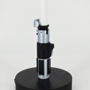 K&ouml;nnte beinhalten: Ein Lichtschwert-Replikat mit einer wei&szlig;en Klinge und einem silbernen und schwarzen Griff. Der Griff weist detaillierte Designelemente auf, darunter Kn&ouml;pfe und Paneele. Das Lichtschwert ist auf einem schwarzen kreisf&ouml;rmigen Sockel montiert.