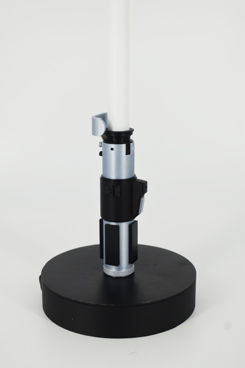 K&ouml;nnte beinhalten: Ein Lichtschwert-Replikat mit einer wei&szlig;en Klinge und einem silbernen und schwarzen Griff. Der Griff weist detaillierte Designelemente auf, darunter Kn&ouml;pfe und Paneele. Das Lichtschwert ist auf einem schwarzen kreisf&ouml;rmigen Sockel montiert.