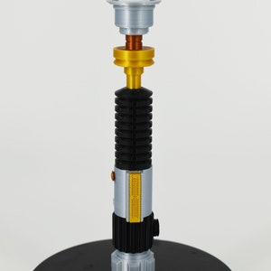 K&ouml;nnte beinhalten: Ein silberner und schwarzer Kerzenhalter in Form eines Lichtschwerts mit einem goldenen Akzentringe. Der Kerzenhalter steht auf einem schwarzen runden Sockel.