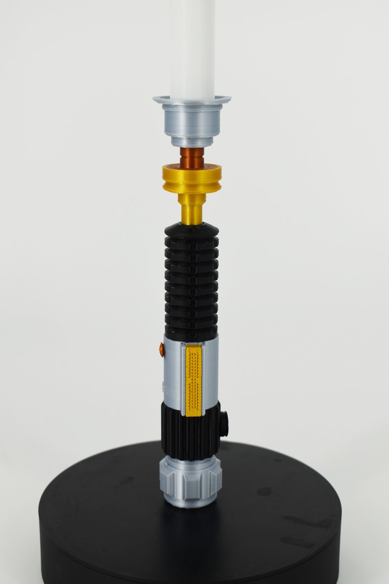 K&ouml;nnte beinhalten: Ein silberner und schwarzer Kerzenhalter in Form eines Lichtschwerts mit einem goldenen Akzentringe. Der Kerzenhalter steht auf einem schwarzen runden Sockel.