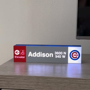 May include: A rectangular sign with the word "Addison" in white text, along with the coordinates "3600 N 940 W". The sign also features the Chicago Cubs logo and an elevator symbol. The sign is on a gray surface.