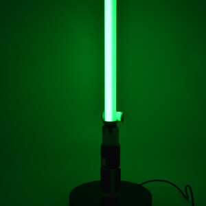 K&ouml;nnte beinhalten: Eine gr&uuml;ne, lichtschwertf&ouml;rmige Lampe mit schwarzem Sockel und Griff. Die Lampe strahlt ein helles gr&uuml;nes Leuchten von der Klinge aus, und der Griff hat schwarze und silberne Details. Die Lampe ist mit einem schwarzen Kabel angeschlossen.