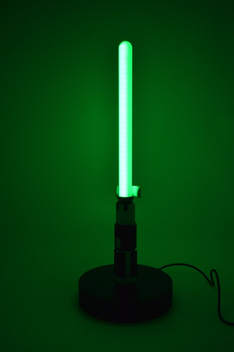 K&ouml;nnte beinhalten: Eine gr&uuml;ne, lichtschwertf&ouml;rmige Lampe mit schwarzem Sockel und Griff. Die Lampe strahlt ein helles gr&uuml;nes Leuchten von der Klinge aus, und der Griff hat schwarze und silberne Details. Die Lampe ist mit einem schwarzen Kabel angeschlossen.